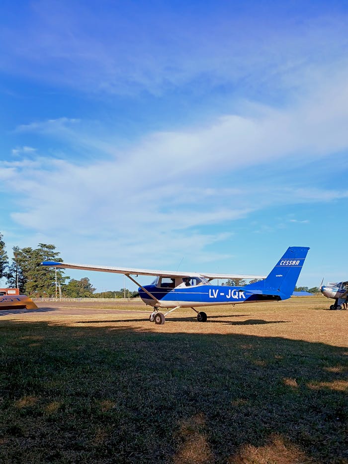 A vibrant blue Cessna airplane parked at a grassy rural airfield under a clear blue sky.